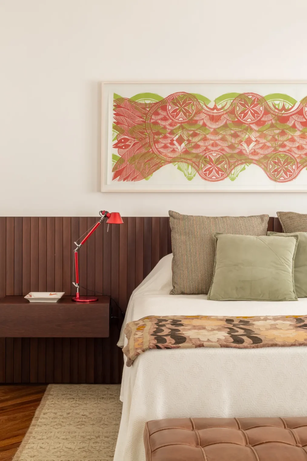 Quarto principal com cabeceira em painel ripado de madeira escura, mesa de cabeceira suspensa com luminária vermelha, cama vestida com almofadas verdes e quadro horizontal estampado na parede clara.