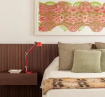 Quarto principal com cabeceira em painel ripado de madeira escura, mesa de cabeceira suspensa com luminária vermelha, cama vestida com almofadas verdes e quadro horizontal estampado na parede clara.