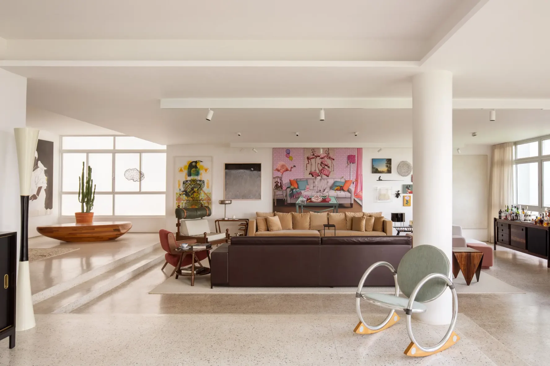 Vista geral da ampla sala de estar integrada com múltiplos ambientes, pilar circular branco, sofás, poltronas de design, banco de madeira e piso de granilite claro.