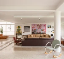 Vista geral da ampla sala de estar integrada com múltiplos ambientes, pilar circular branco, sofás, poltronas de design, banco de madeira e piso de granilite claro.