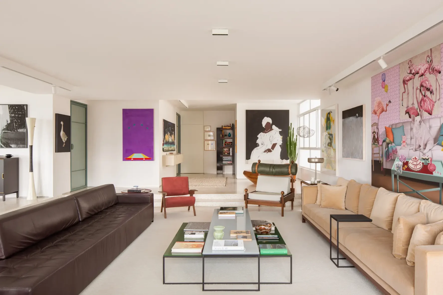 Texto Alternativo: Perspectiva longitudinal da sala de estar com sofá de couro escuro, sofá bege, mesa de centro dupla com livros, poltronas de design e vista para o hall de entrada ao fundo sobre piso de granilite.