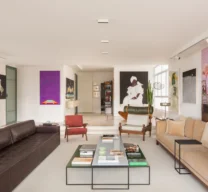 Texto Alternativo: Perspectiva longitudinal da sala de estar com sofá de couro escuro, sofá bege, mesa de centro dupla com livros, poltronas de design e vista para o hall de entrada ao fundo sobre piso de granilite.