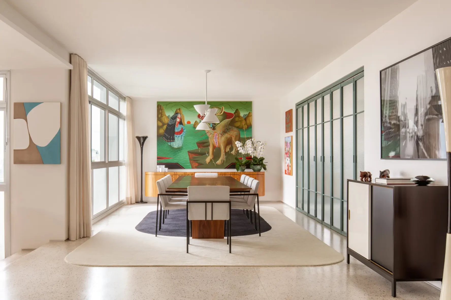 Sala de jantar ampla com mesa de madeira, cadeiras estofadas claras sobre tapete claro com centro escuro, buffet curvo com pintura verde de fundo e painéis em serralheria verde à direita.