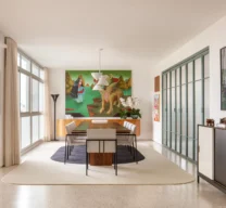 Sala de jantar ampla com mesa de madeira, cadeiras estofadas claras sobre tapete claro com centro escuro, buffet curvo com pintura verde de fundo e painéis em serralheria verde à direita.