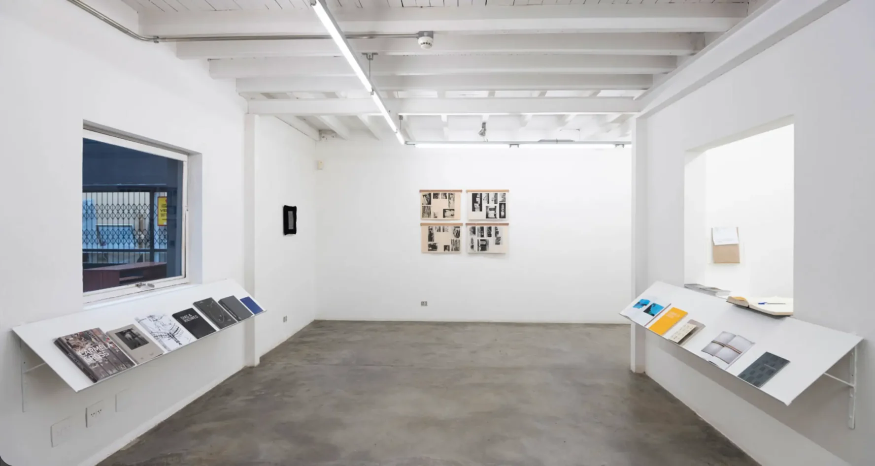 Texto Alternativo: Sala de exposição de galeria de arte com paredes brancas, piso de cimento queimado, teto com vigas expostas e prateleiras inclinadas expondo livros e catálogos.