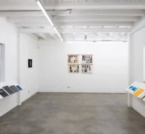 Texto Alternativo: Sala de exposição de galeria de arte com paredes brancas, piso de cimento queimado, teto com vigas expostas e prateleiras inclinadas expondo livros e catálogos.