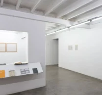 Vista interna da Galeria Vermelho mostrando abertura na parede com expositor inclinado de livros, iluminação tubular e quadros emoldurados ao fundo.