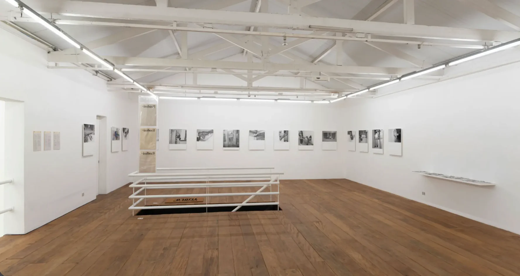 Sala de exposição superior na Galeria Vermelho com piso de assoalho de madeira, guarda-corpo branco, vigas de telhado expostas e série de fotografias em preto e branco, de Marcelo Cidade, nas paredes.