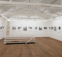 Sala de exposição superior na Galeria Vermelho com piso de assoalho de madeira, guarda-corpo branco, vigas de telhado expostas e série de fotografias em preto e branco, de Marcelo Cidade, nas paredes.