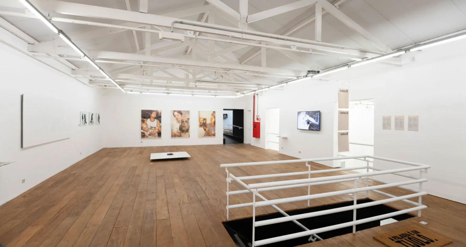 Pavimento superior da galeria de arte com piso de tábuas de madeira, tesouras de telhado expostas em branco, guarda-corpo de metal e obras de arte dispostas nas paredes.