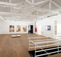 Pavimento superior da galeria de arte com piso de tábuas de madeira, tesouras de telhado expostas em branco, guarda-corpo de metal e obras de arte dispostas nas paredes.