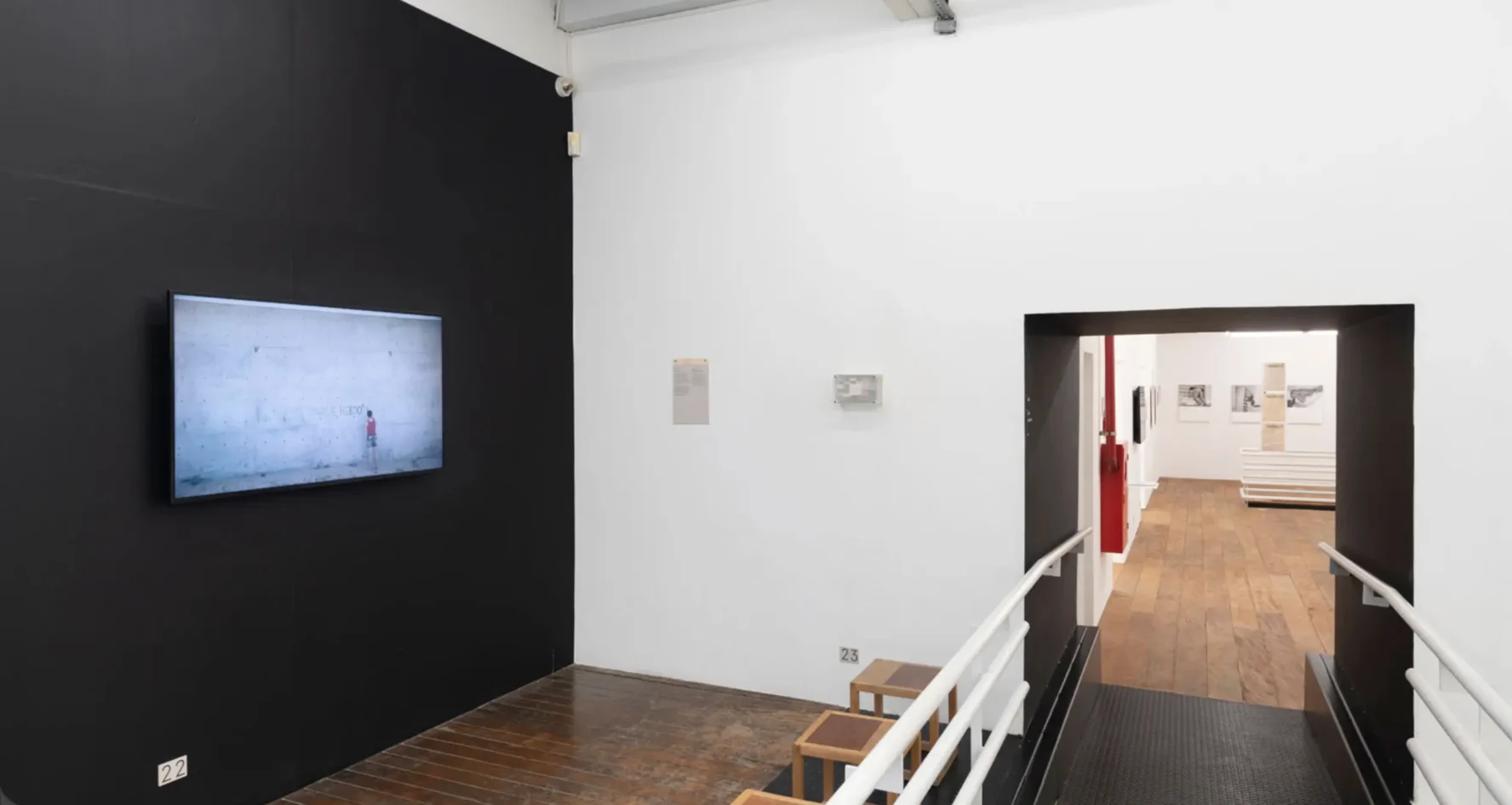 Sala de videoarte na Galeria Vermelho com parede preta, tela de exibição, rampa de acesso em chapa metálica com corrimão branco e passagem para a sala adjacente com piso de madeira.