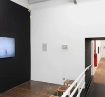 Sala de videoarte na Galeria Vermelho com parede preta, tela de exibição, rampa de acesso em chapa metálica com corrimão branco e passagem para a sala adjacente com piso de madeira.