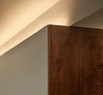 Detalhe de iluminação indireta embutida na sanca do teto sobre o encontro de parede clara e painel de madeira escura no Edifício Flor de Liz.