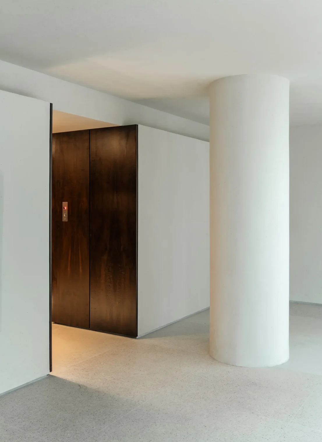 Hall dos elevadores do Edifício Flor de Liz com coluna circular em concreto claro, nicho em marcenaria de madeira escura e piso claro.
