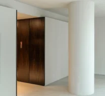 Hall dos elevadores do Edifício Flor de Liz com coluna circular em concreto claro, nicho em marcenaria de madeira escura e piso claro.