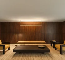 Enquadramento frontal da sala de estar do Edifício Flor de Liz com duas poltronas amarelas, banco estofado, mesa de centro baixa, luminária pendente e paredes em madeira escura.