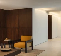 Transição entre a sala de estar revestida em madeira com poltrona amarela e o corredor amplo iluminado com coluna circular no Edifício Flor de Liz.