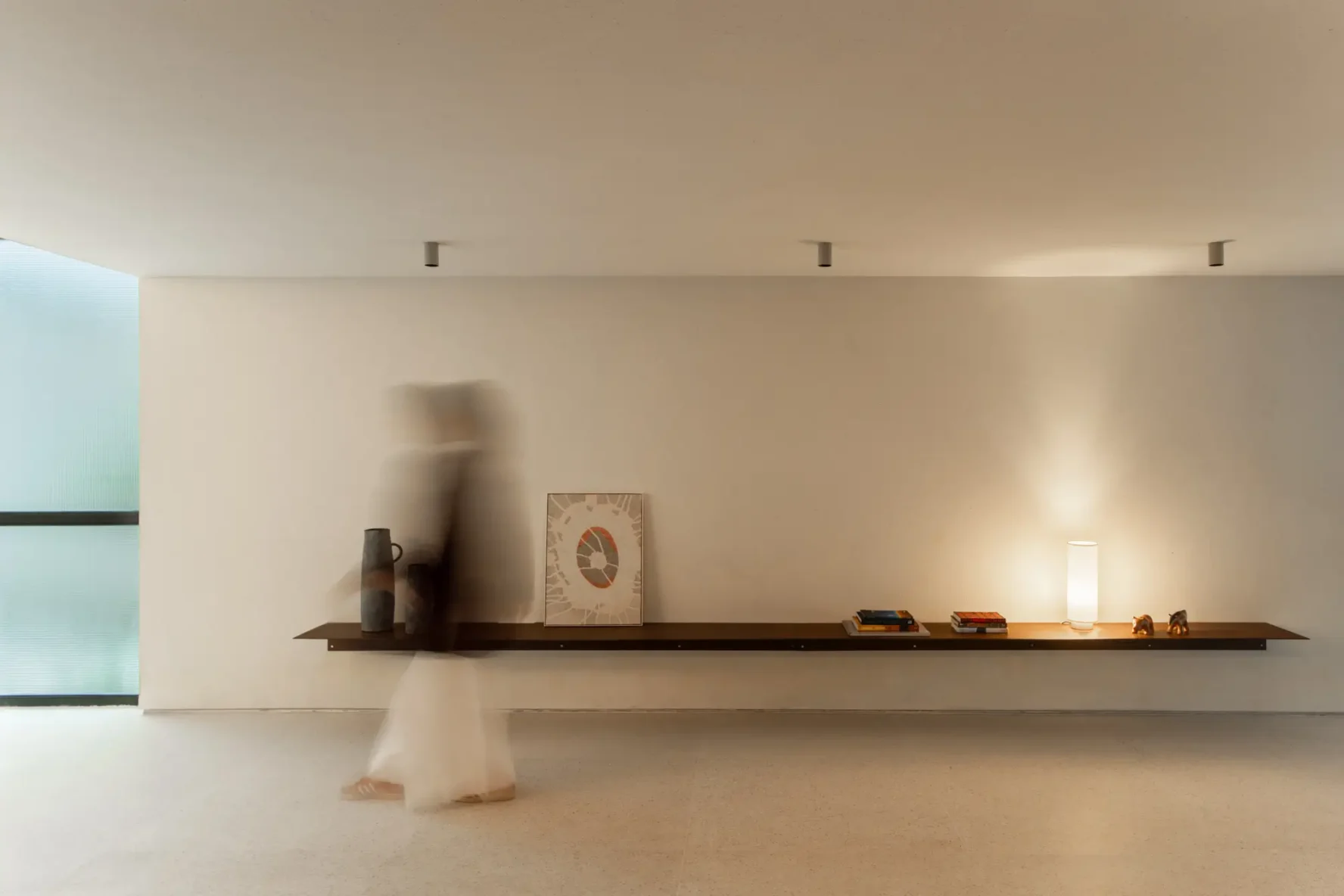 Prateleira metálica contínua suspensa em parede clara com iluminação, objetos decorativos e figura humana desfocada caminhando no corredor do Edifício Flor de Liz.