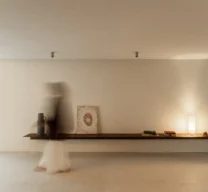 Prateleira metálica contínua suspensa em parede clara com iluminação, objetos decorativos e figura humana desfocada caminhando no corredor do Edifício Flor de Liz.