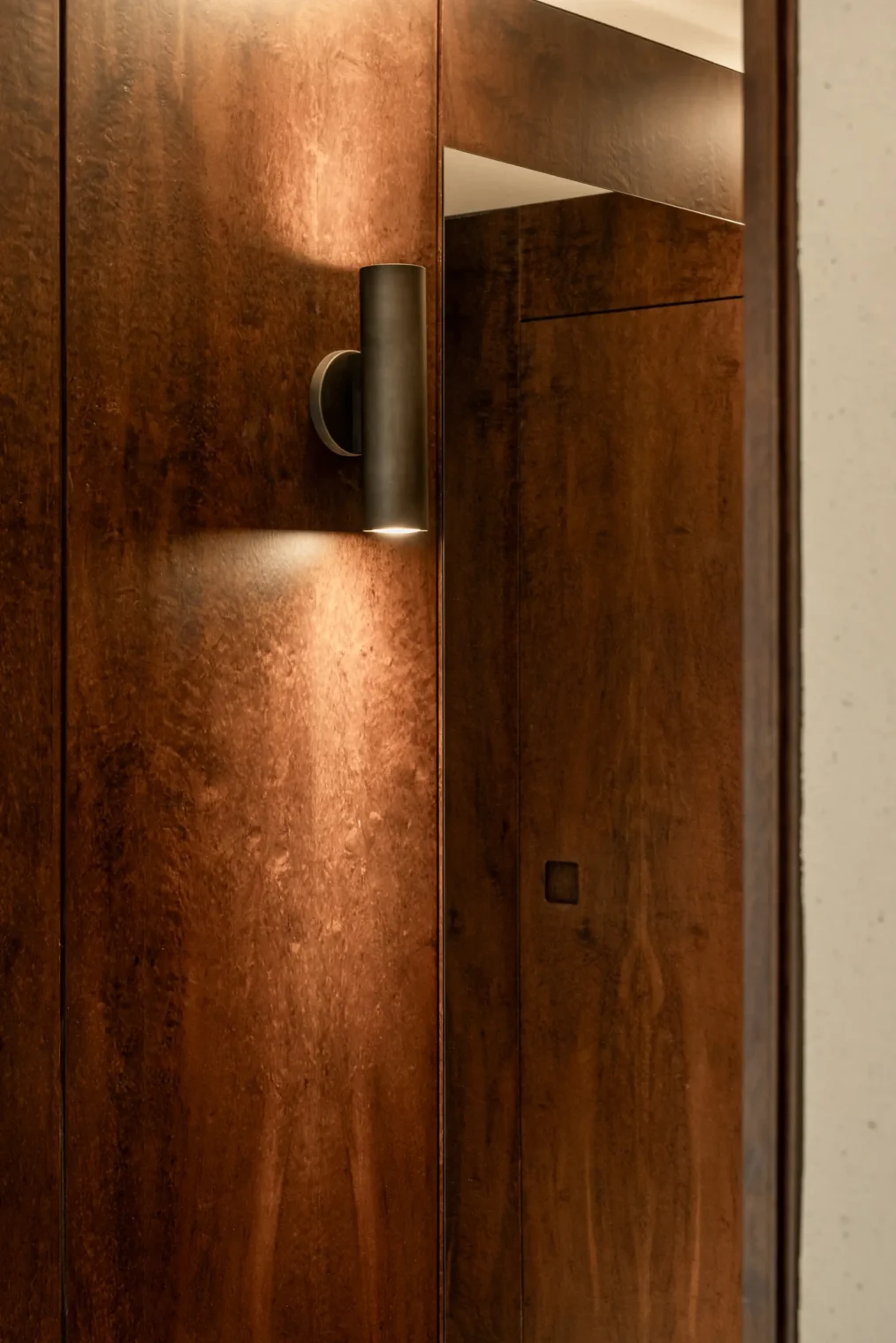 Arandela cilíndrica de luz bidirecional sobre painel de madeira escura com reflexo de porta mimetizada em espelho no Edifício Flor de Liz.