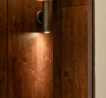 Arandela cilíndrica de luz bidirecional sobre painel de madeira escura com reflexo de porta mimetizada em espelho no Edifício Flor de Liz.