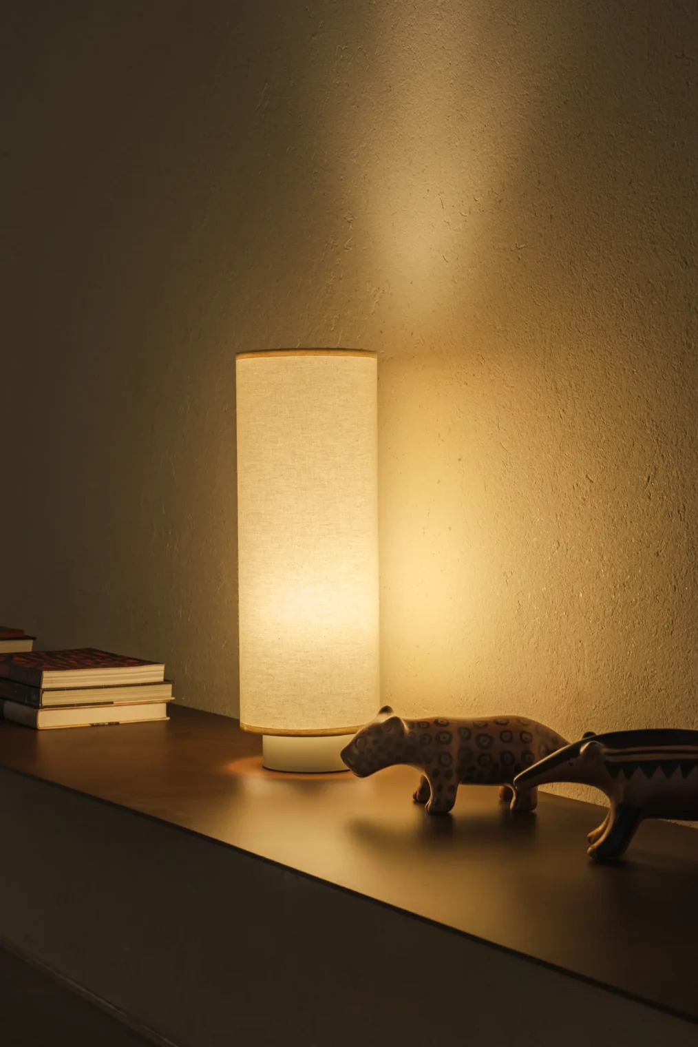 Luminária de mesa cilíndrica de linho acesa sobre prateleira de madeira com esculturas de animais e livros contra parede texturizada no Edifício Flor de Liz.