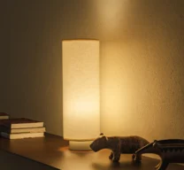 Luminária de mesa cilíndrica de linho acesa sobre prateleira de madeira com esculturas de animais e livros contra parede texturizada no Edifício Flor de Liz.