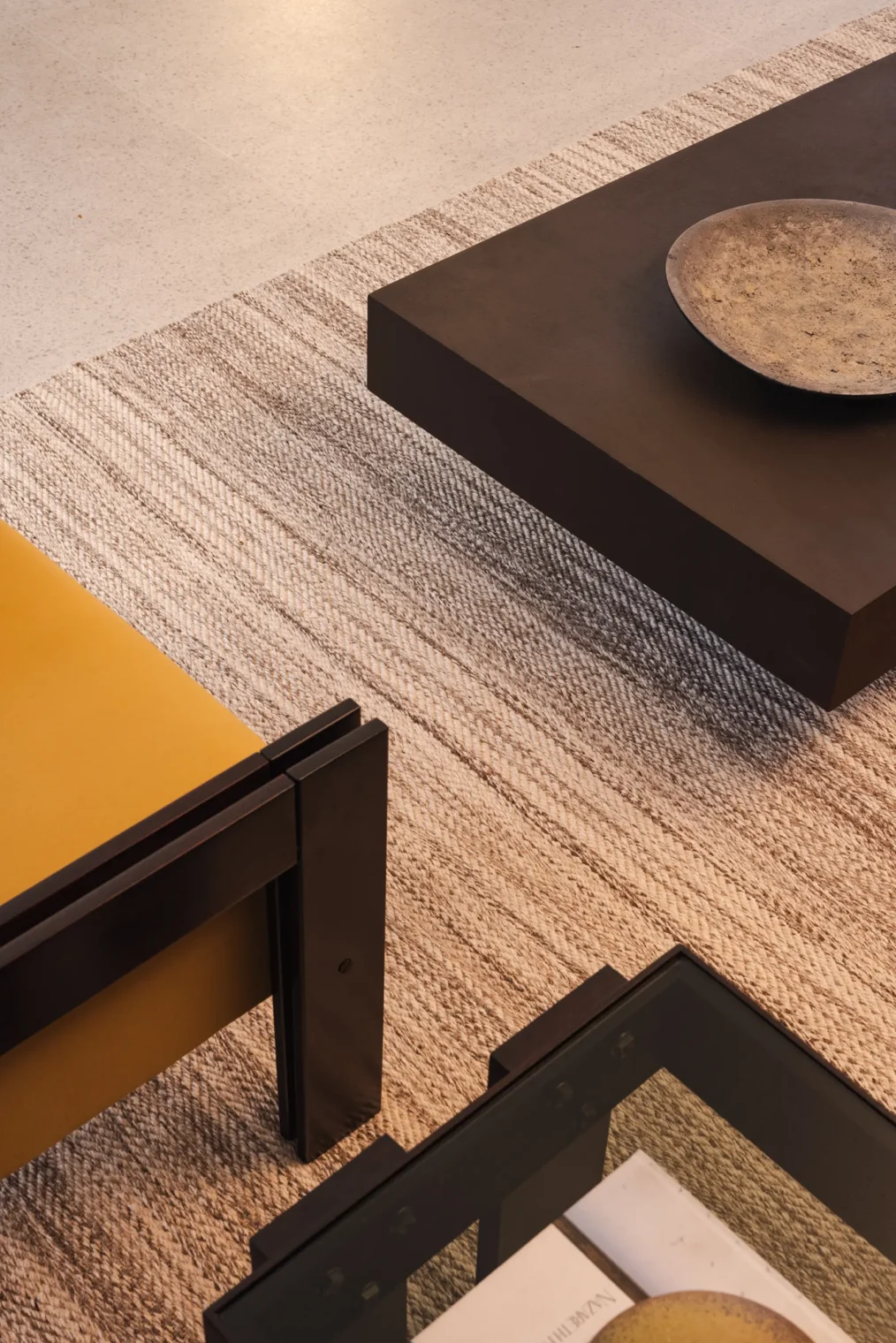 Enquadramento superior e detalhe das texturas no estar do Edifício Flor de Liz com prato cerâmico sobre mesa de centro escura, estofado amarelo, mesa com tampo de vidro e tapete de fibra natural.