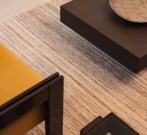 Enquadramento superior e detalhe das texturas no estar do Edifício Flor de Liz com prato cerâmico sobre mesa de centro escura, estofado amarelo, mesa com tampo de vidro e tapete de fibra natural.