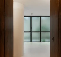 Moldura de painéis em madeira escura abrindo vista para pilar circular claro e divisória de vidro aramado com caixilharia verde no Edifício Flor de Liz.