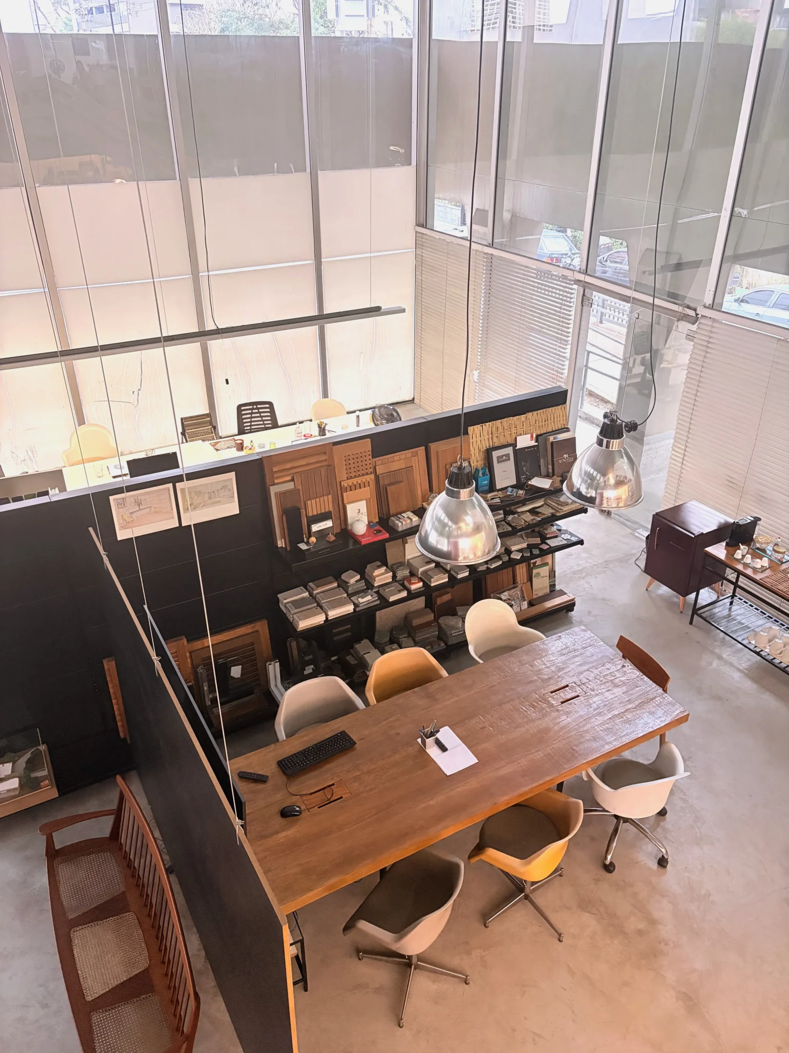 Vista aérea de escritório de arquitetura com pé-direito duplo, mesa de reunião em madeira rústica com pendentes metálicos, estante preta com amostras de materiais e estações de trabalho ao fundo.